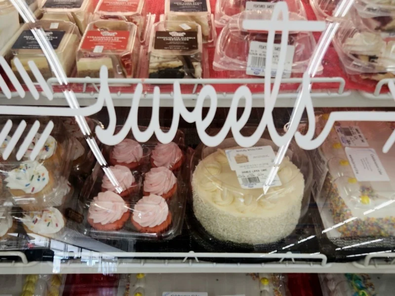 Bakery case of frosted cupcakes and a whole cake under the Sweets sign
