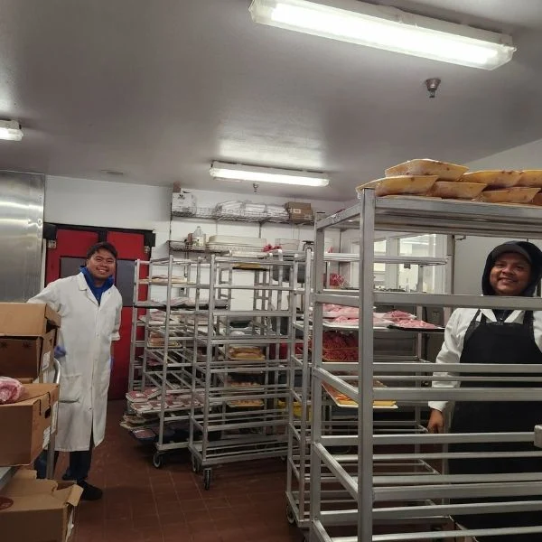 Two Olsens Marketplace kitchen employees beside bakery racks